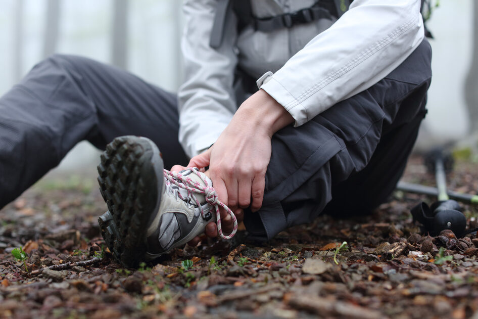 A person dressed in outdoor clothing sits on the ground in the forest, holding one ankle with both hands.