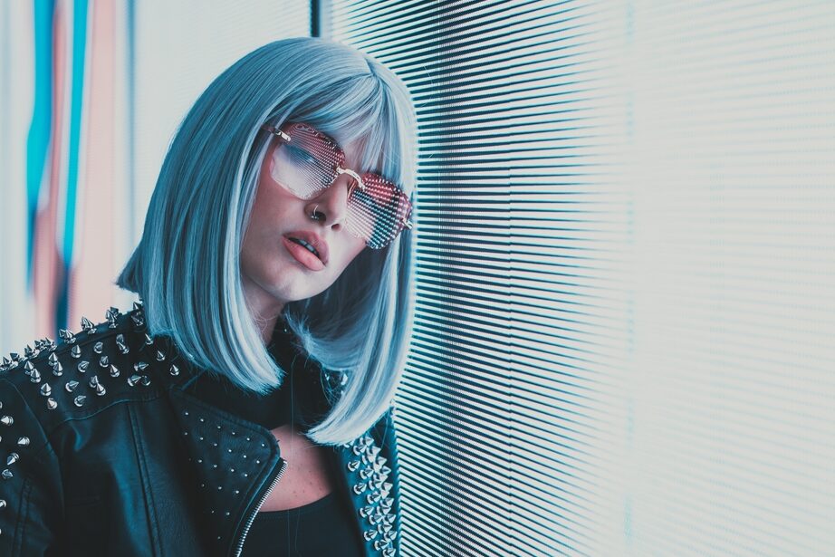 A person with a turquoise wig stands confidently in pastel-colored lighting.