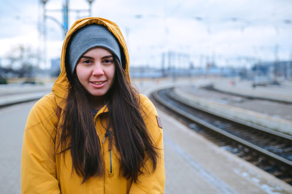 A young woman wearing a beanie stands on a train station platform, looking straight ahead.