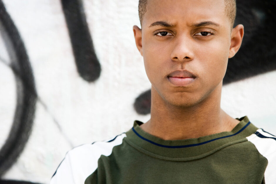 A young man stands outdoors by a spray-painted wall, staring ahead with a serious, furrowed-brow expression.