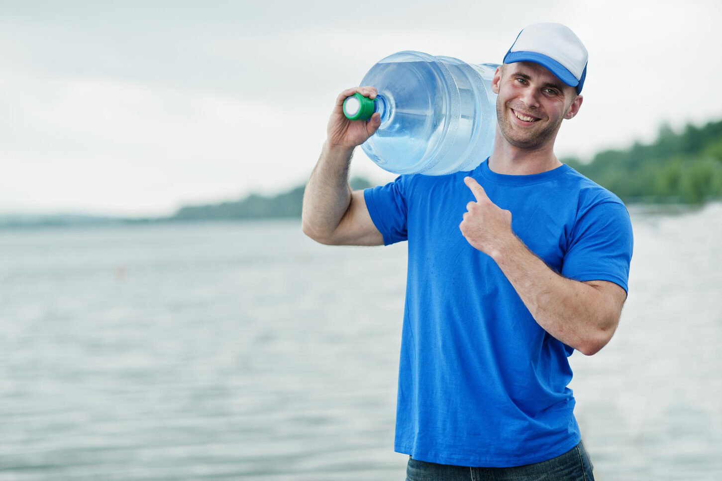 A smiling young man carries a large water container on his shoulder. A shoreline and body of water are in the background.