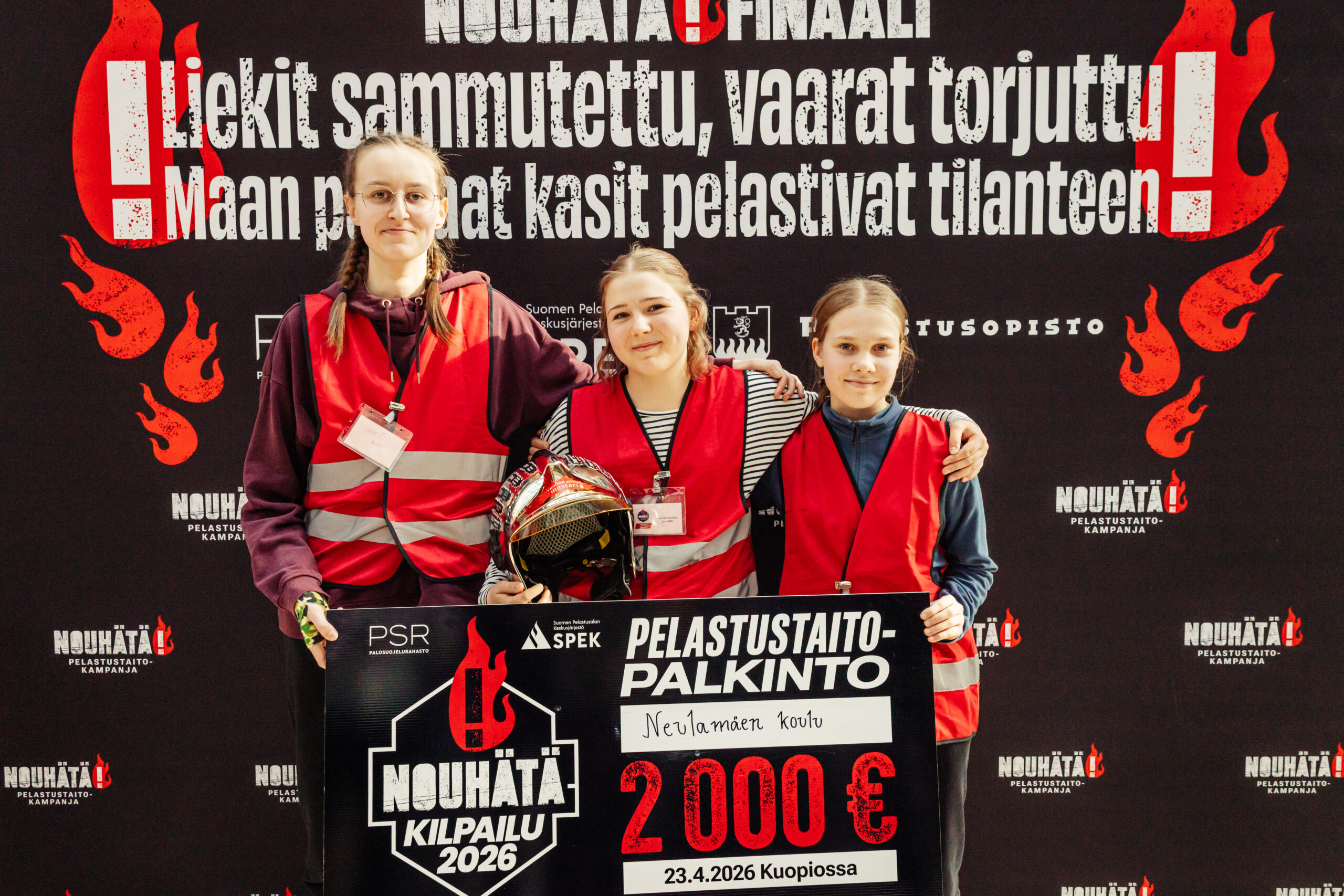 NouHätä-kilpailun voittaneet kolme tyttöä seisovat hymyillen rivissä palkintosekin kanssa.