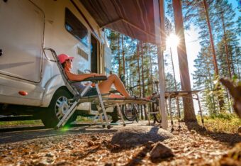 Nainen istuu aurinkotuolissa matkailuauton edessä metsäisellä leirintäalueella.