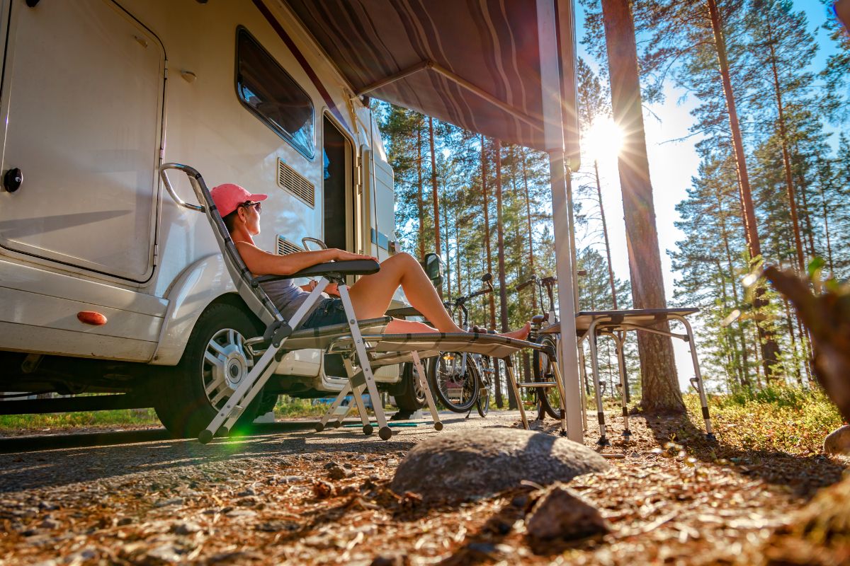 Nainen istuu aurinkotuolissa matkailuauton edessä metsäisellä leirintäalueella.