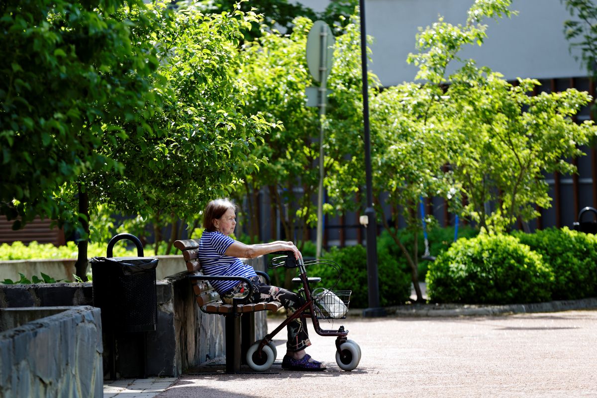Vanhempi nainen istuu puiston penkillä rollaattorin kanssa.